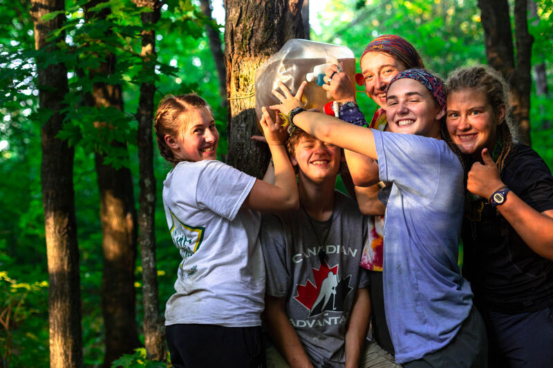 A group of five young people are gathered around a tree in a wooded area. One person is holding a large, clear container against the tree. The others are smiling and looking at the container. They appear to be enjoying an outdoor activity or project together in a natural setting. The scene is bathed in sunlight filtering through the trees.